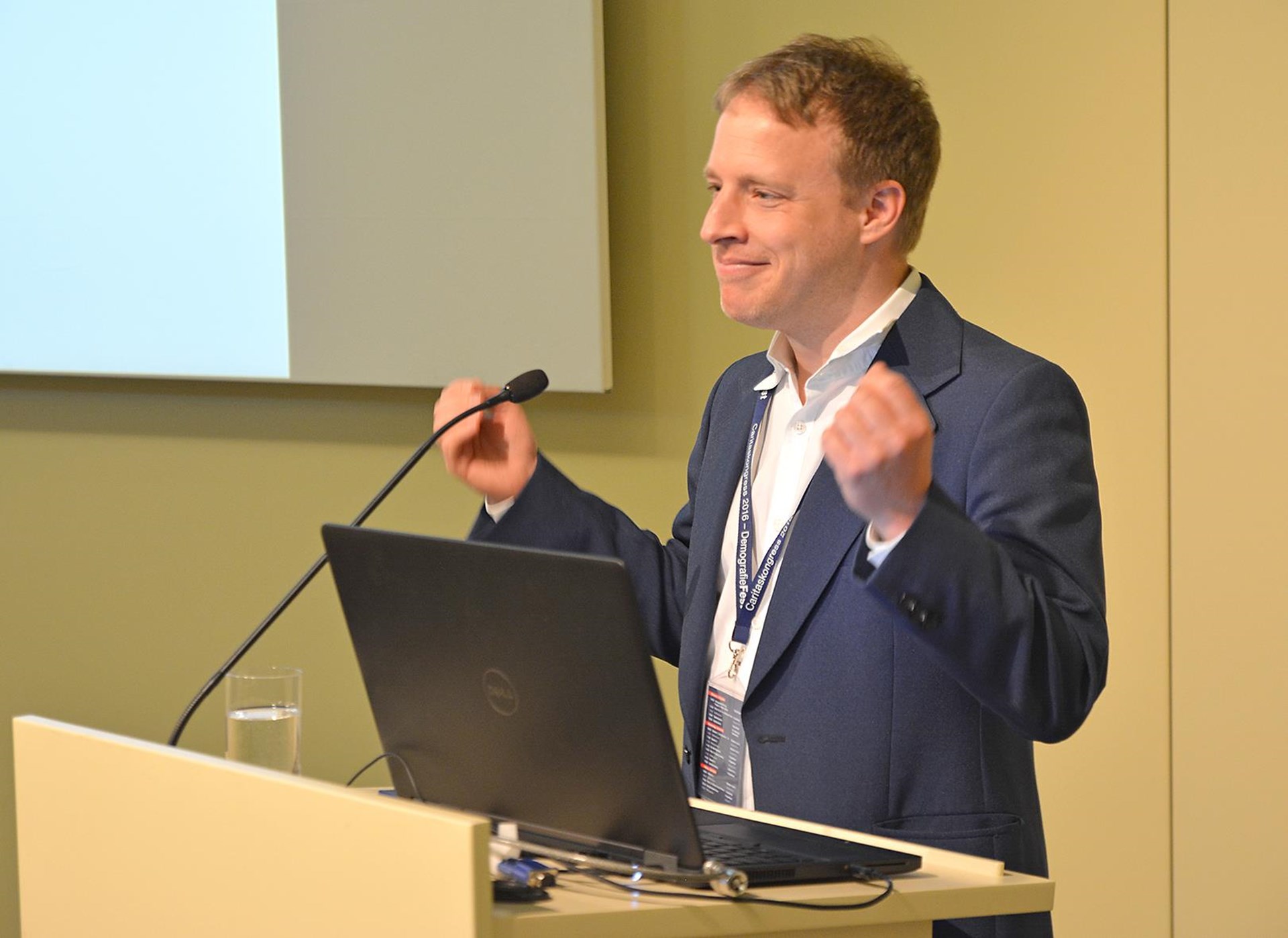 Mann in Anzug spricht an einem Podium, lächelt und gestikuliert enthusiastisch während einer Präsentation.