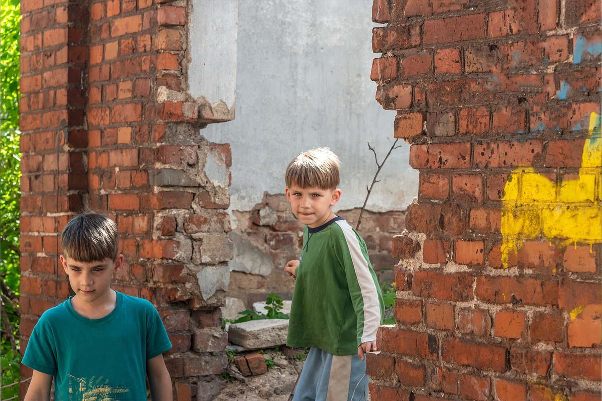 Zwei Jungen in zerrissenen T-Shirts stehen zwischen den Ruinen eines alten Ziegelgebäudes, umgeben von grüner Vegetation.