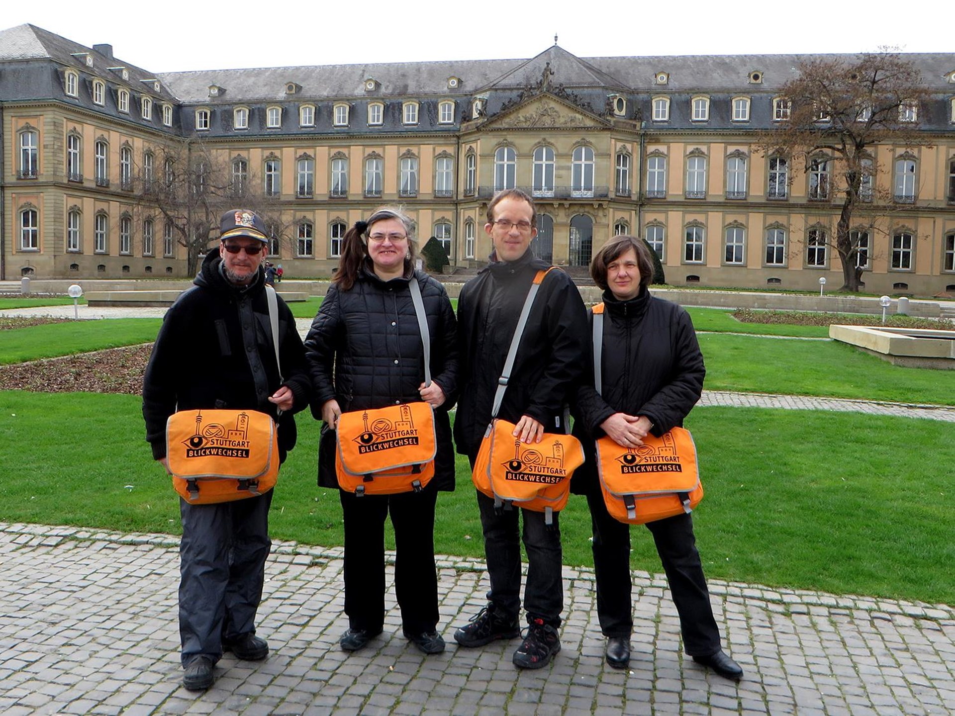Vier Personen stehen vor einem historischen Gebäude, tragen schwarze Kleidung und orangefarbene Umhängetaschen mit dem Aufdruck 'Friedensbotschafter'.