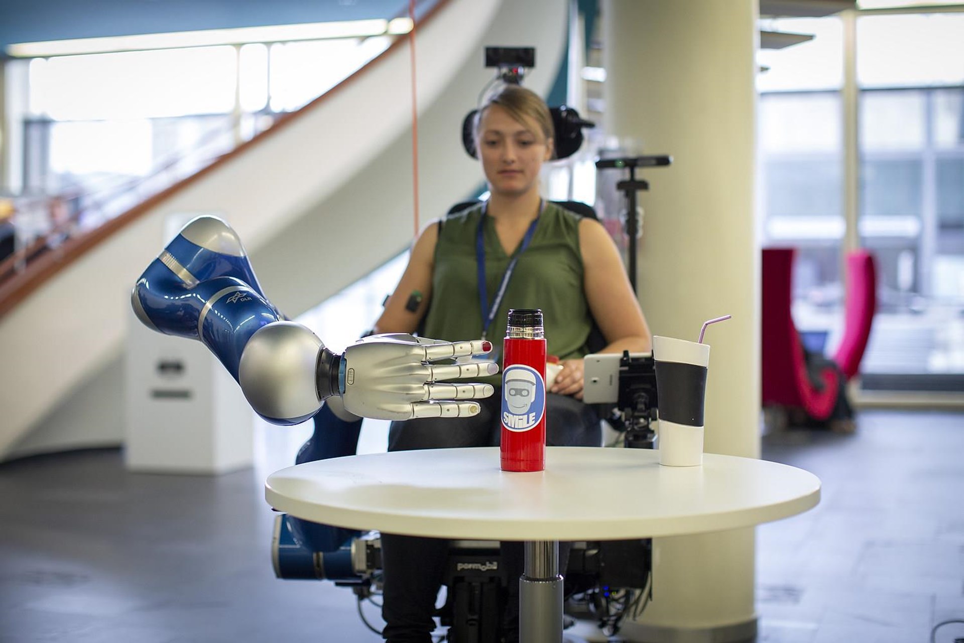 Ein humanoider Roboterarm greift nach einer roten Wasserflasche auf einem Tisch, während eine Person im Hintergrund sitzt und den Roboter steuert.