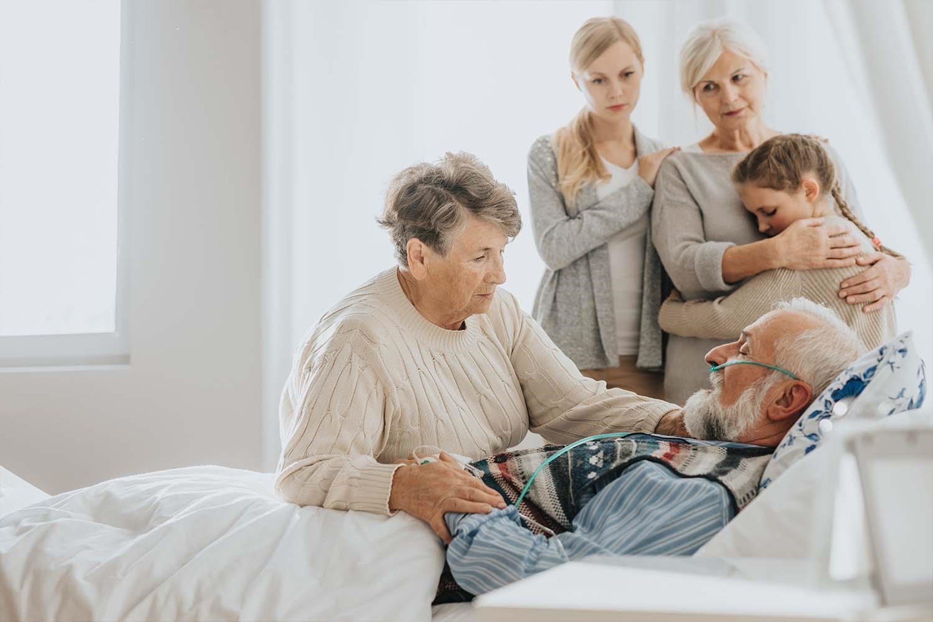 Älterer Mann im Bett mit Sauerstoffmasken, umgeben von drei Frauen, die besorgt und liebevoll mit ihm sprechen. 