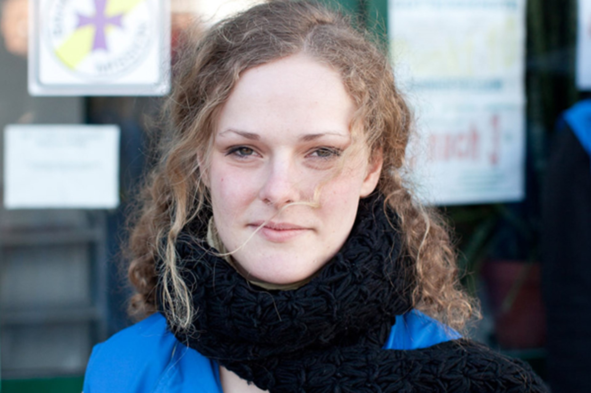 Porträt einer jungen Frau mit lockigem Haar, die einen schwarzen Schal und ein blaues Oberteil trägt. Sie schaut neutral in die Kamera vor einem unscharfen Hintergrund mit Beschilderung.