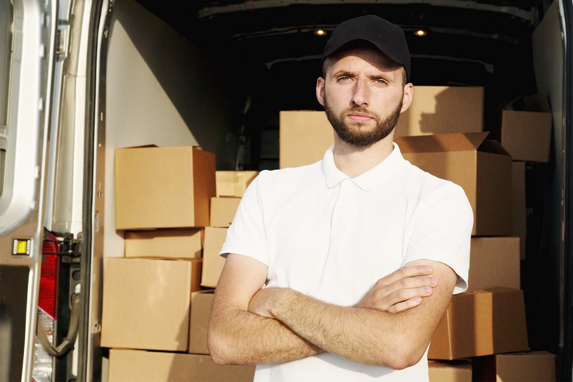 Mann mit Bart steht vor einem Lieferwagen, im Hintergrund sind Kartons gestapelt. Er trägt ein weißes T-Shirt und eine schwarze Kappe, mit verschränkten Armen und ernstem Blick.
