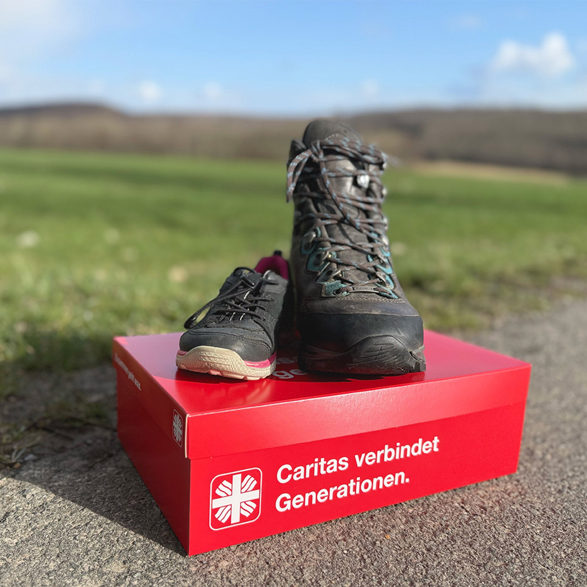 Auf einem Wanderweg im ländlichen Raum steht ein rotrn Kampagnenkarton mit der Aufschrift "Caritas verbindet Generationen!". Darauf ein großer und ein kleiner Wanderschuh.