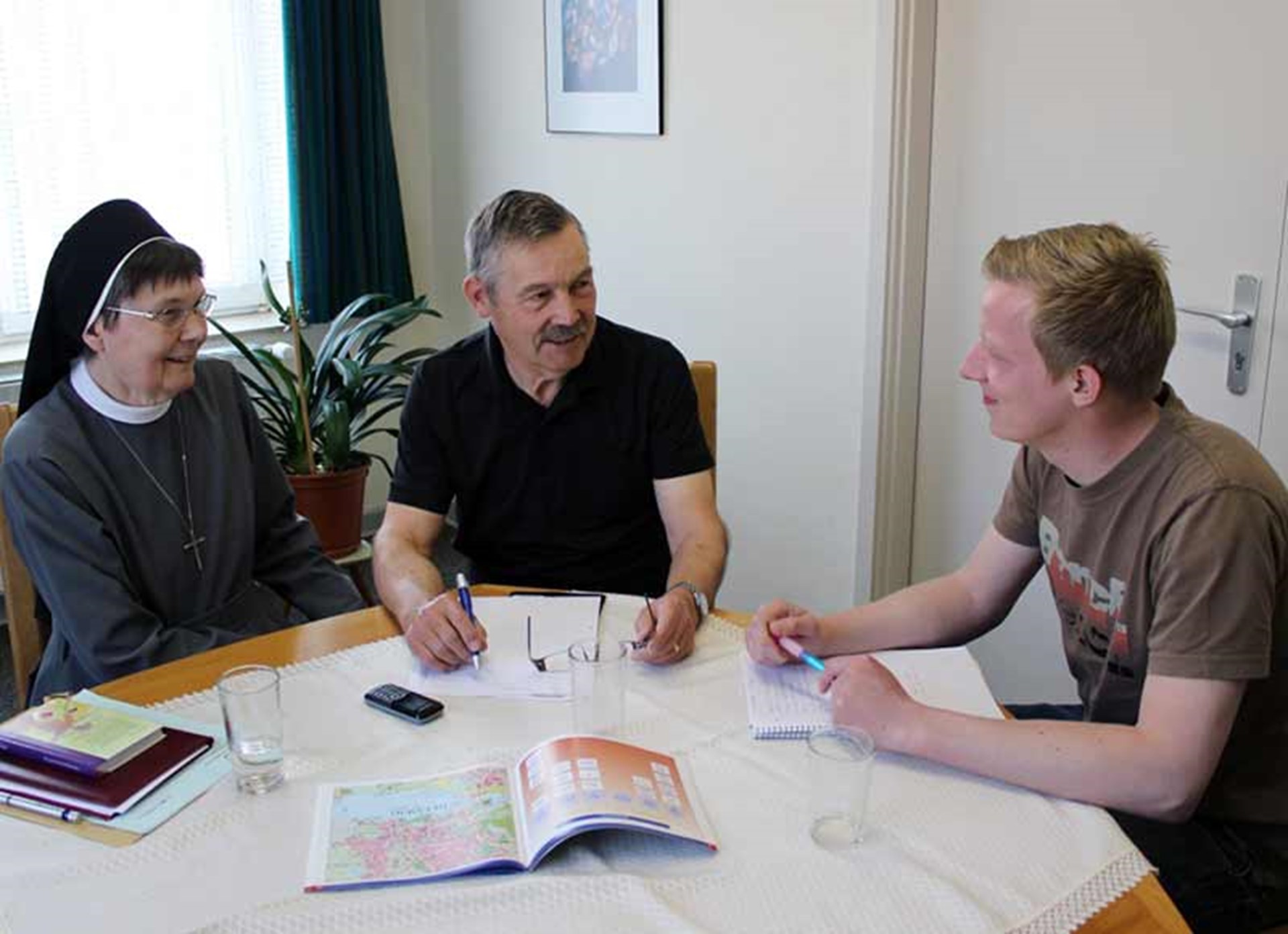 Drei Personen sitzen an einem Tisch und unterhalten sich, darunter eine Nonne, ein älterer Mann und ein junger Mann mit Schreibmaterialien.