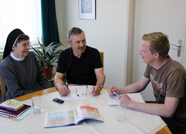 Drei Personen sitzen an einem Tisch und unterhalten sich, darunter eine Nonne, ein älterer Mann und ein junger Mann mit Schreibmaterialien.