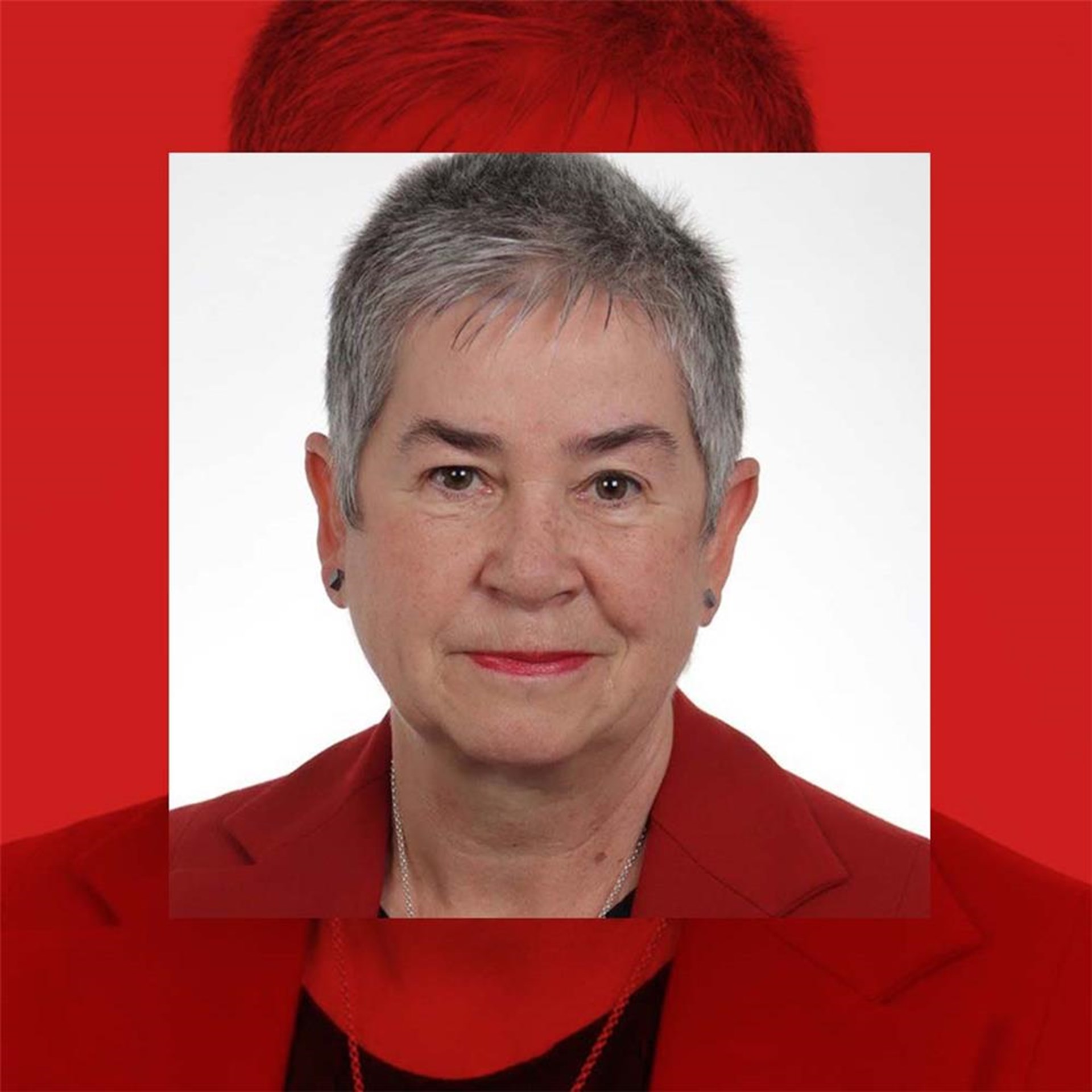 Porträt einer älteren Frau mit grauen Haaren, die einen roten Blazer trägt. Sie hat einen neutralen Gesichtsausdruck und blickt direkt in die Kamera.