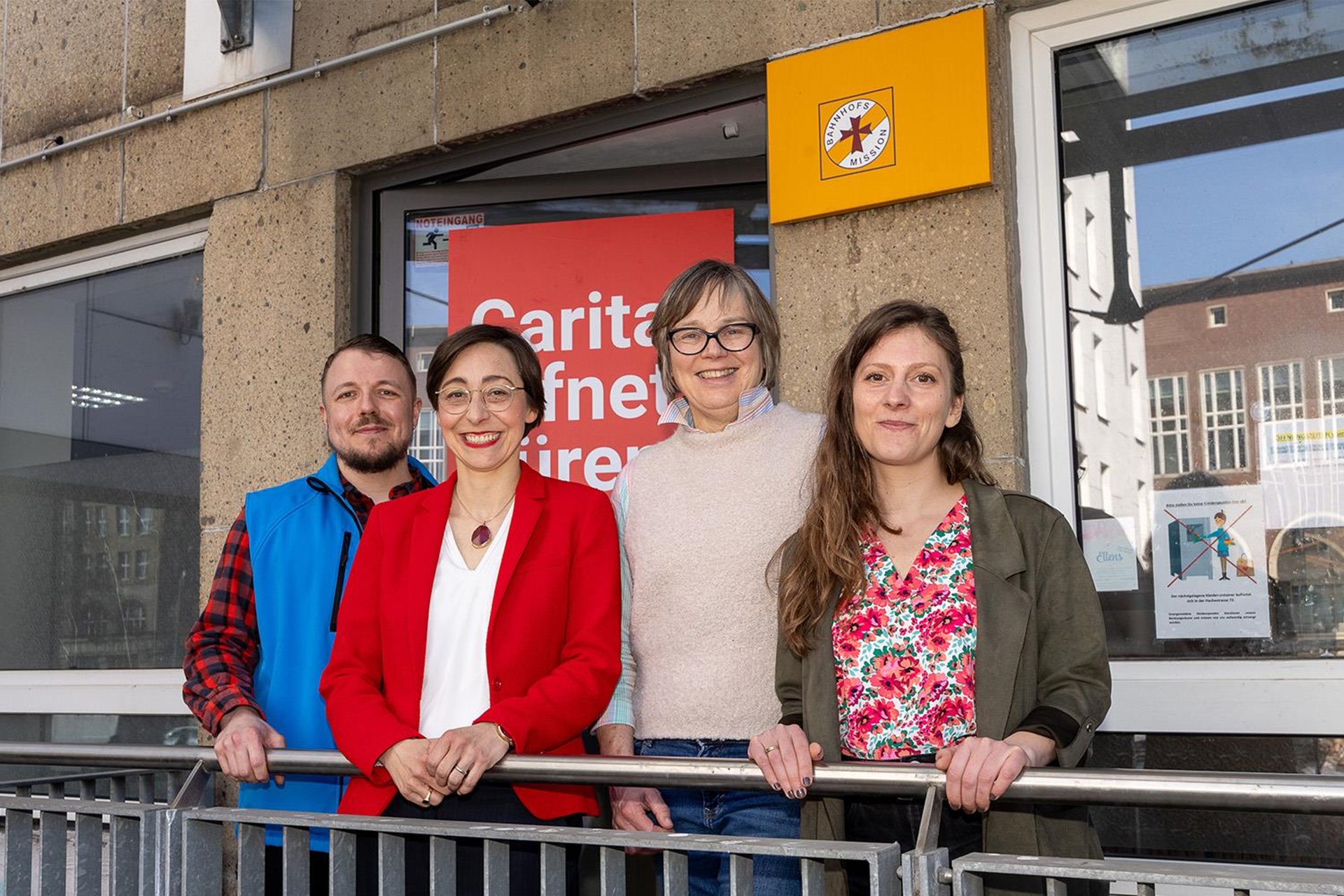 Gruppenbild vor einem Gebäude der Bahnhofsmission, ein Mann und drei Frauen, darunter Caritas-Präsidentin Welskop-Deffaa, hinter ihnen ein rotes Kampagnenplakat "Caritas öffnet Türen".