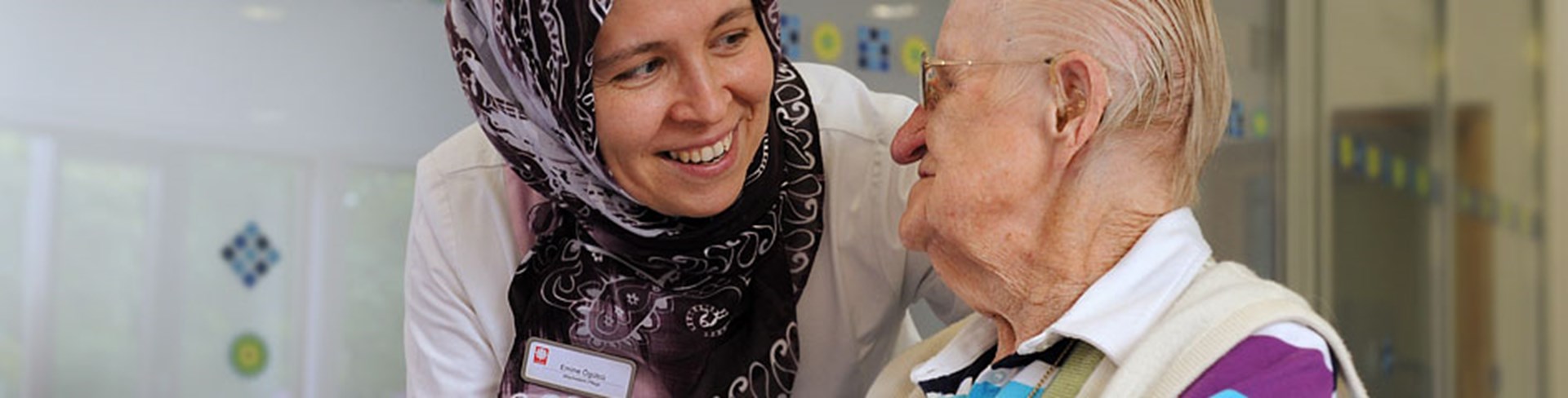 Pflegekraft mit Kopftuch lächelt und spricht mit einer älteren Dame im Pflegeheim.