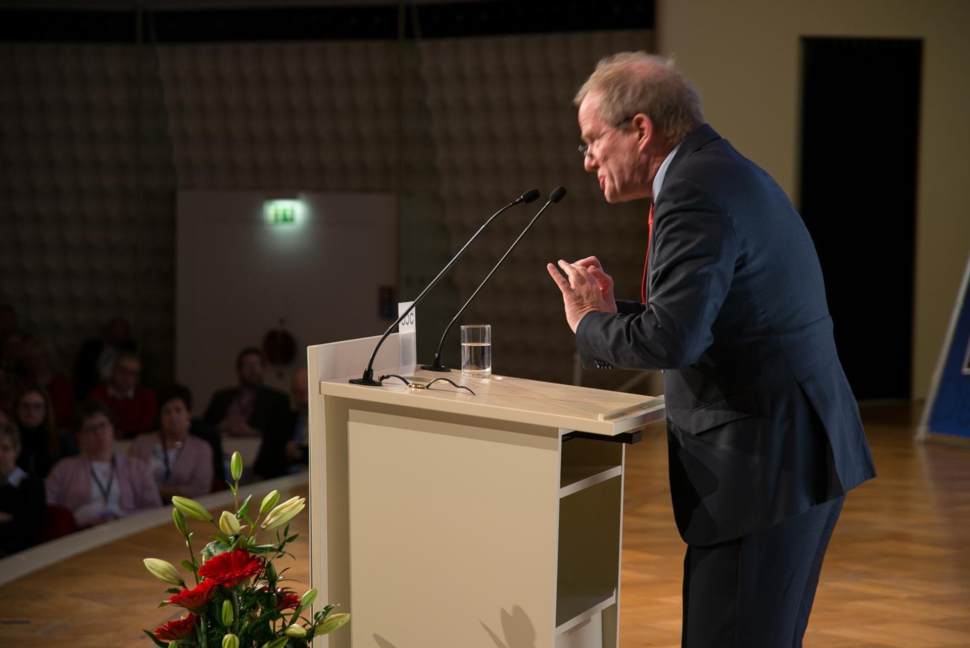 Ein älterer Mann in einem Anzug spricht leidenschaftlich an einem Rednerpult, während eine Gruppe von Zuhörern im Hintergrund sitzt.