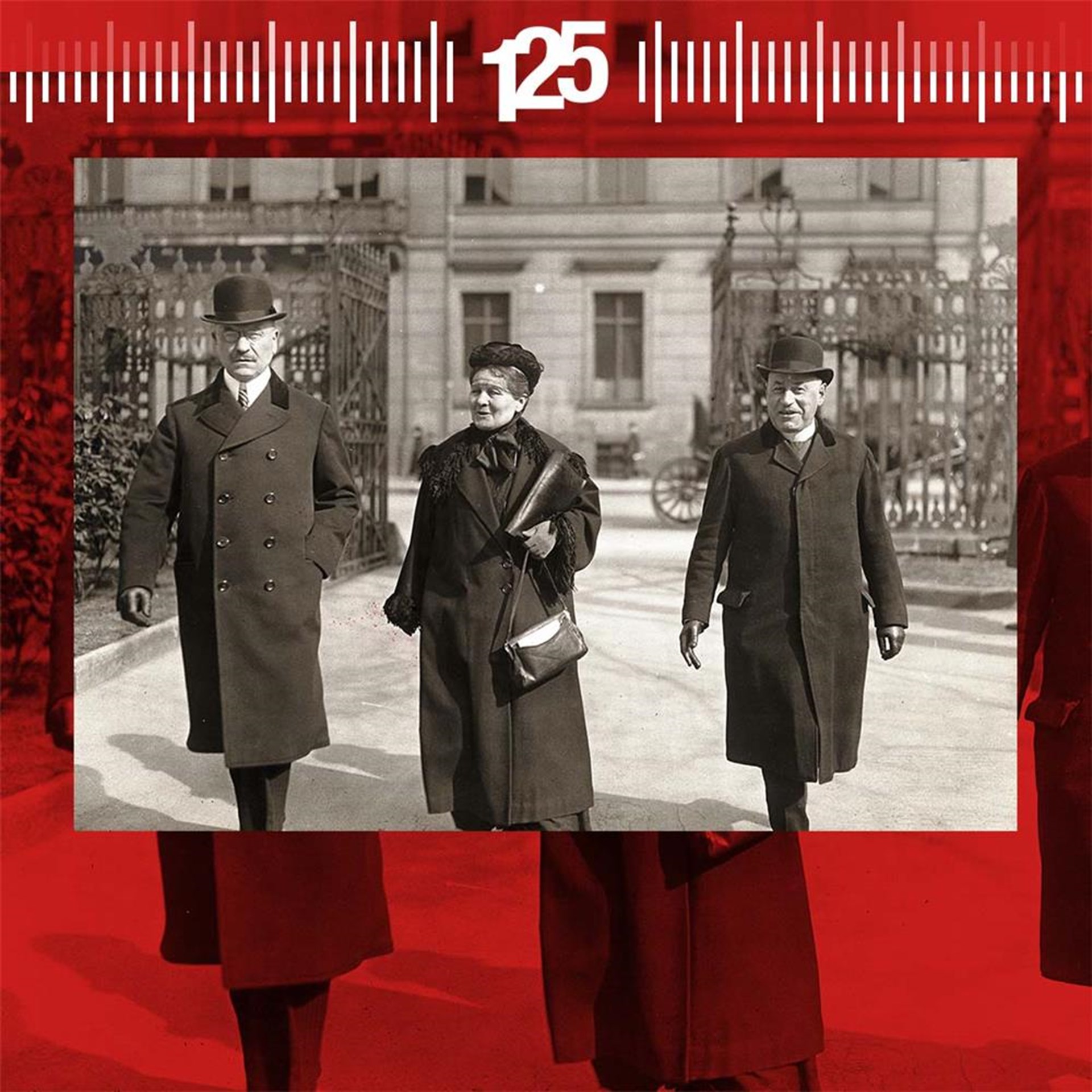 Drei Personen in historischen Anzügen und Hüten gehen auf einer Straße, umgeben von Gebäuden und einem Zaun.