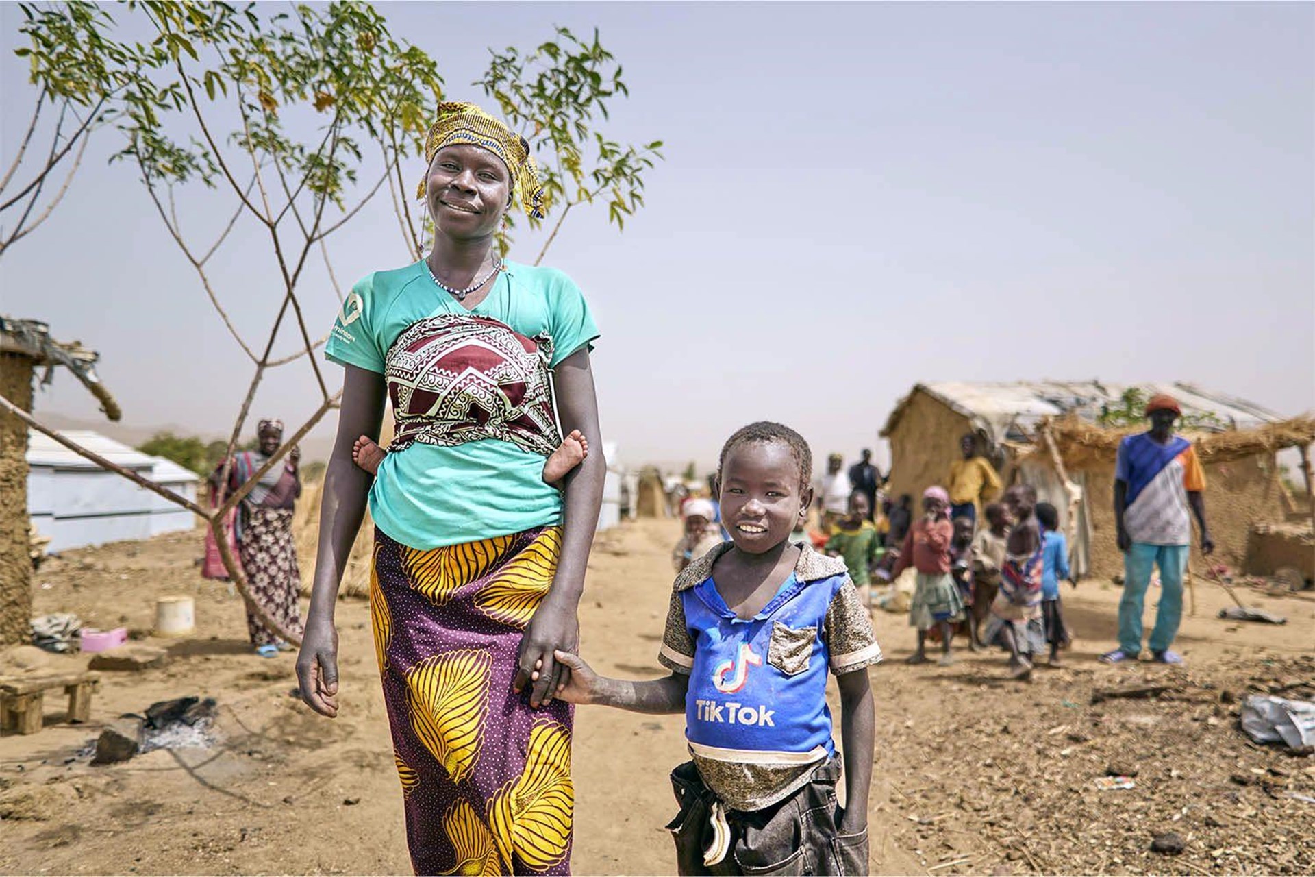 Eine junge bunt gekleidete Frau in Kamerun hält ihren fünfjährigen Sohn an der Hand. Im Hintergrund sind Strohhütten auf trockenem Gelände zu sehen.