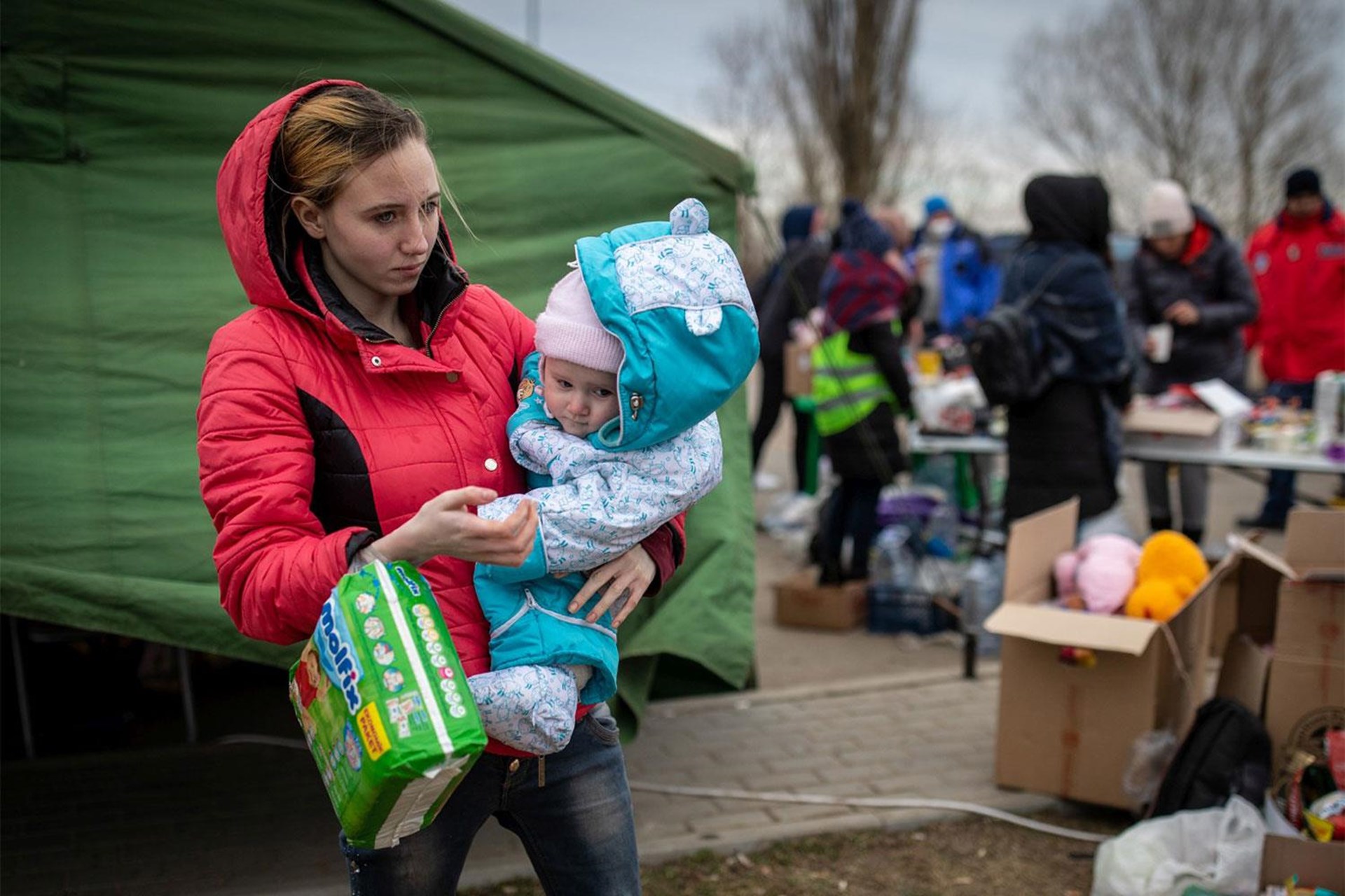 Eine Frau in roter Jacke hält ein Baby in blauer Winterjacke und trägt eine Packung Windeln. Im Hintergrund sind Menschen und Tische mit Hilfsgütern.