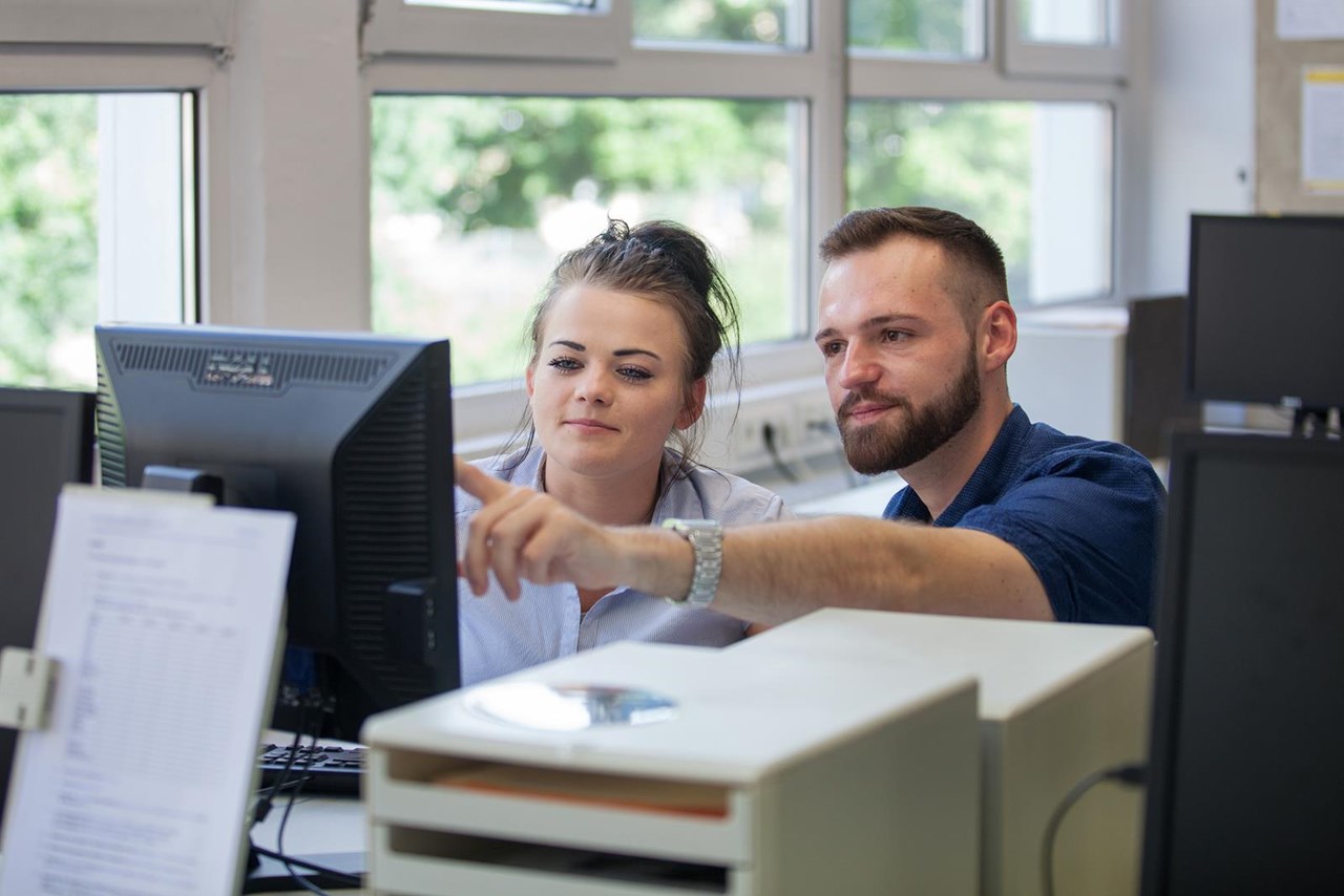 Ein Mann und eine Frau sitzen an einem Schreibtisch und schauen auf einen Computerbildschirm. Der Mann zeigt mit dem Finger auf den Bildschirm, während die Frau aufmerksam zuschaut.