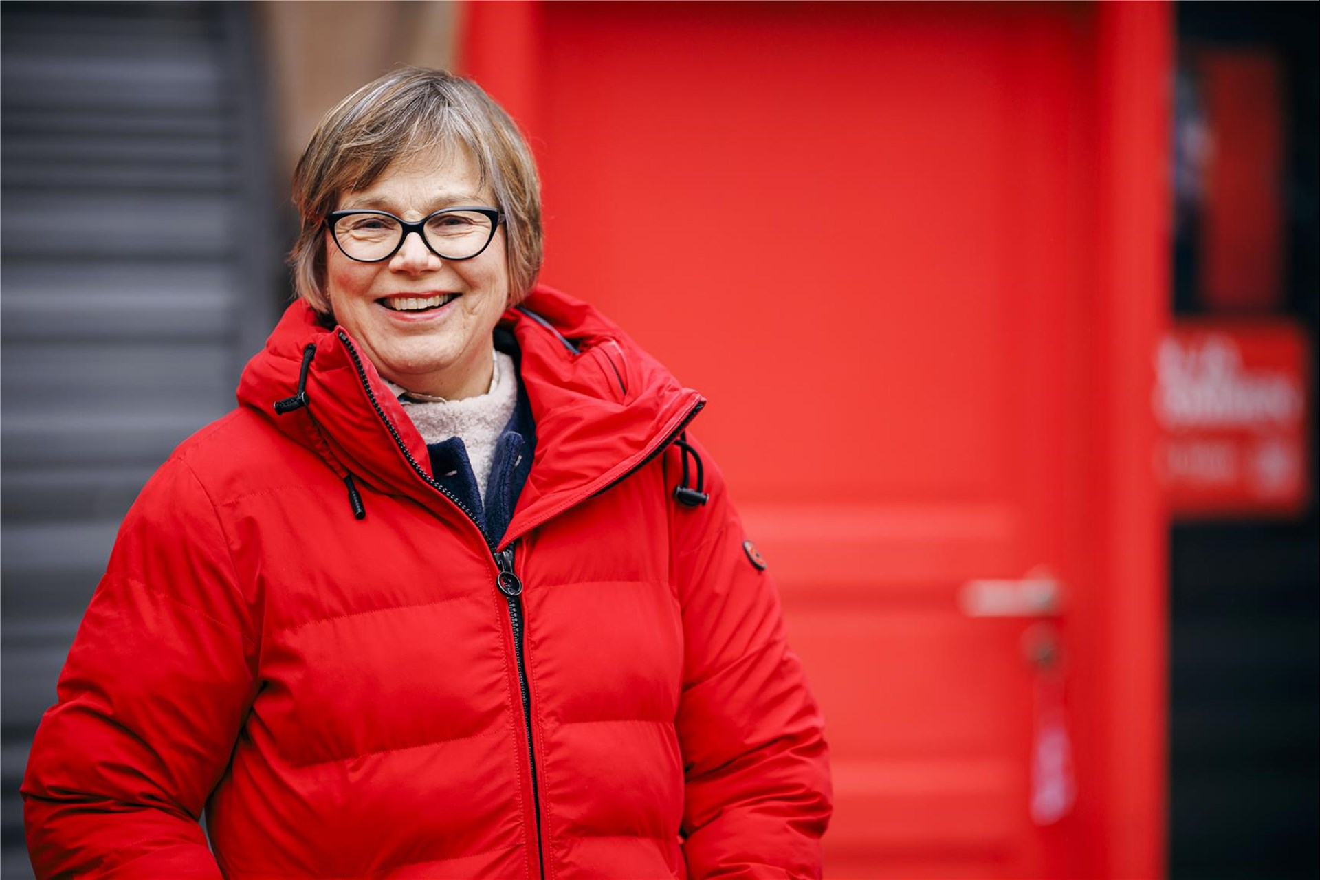 Eine lächelnde Frau mit kurzen, braunen Haaren und Brille trägt eine rote Winterjacke vor einer roten Tür.