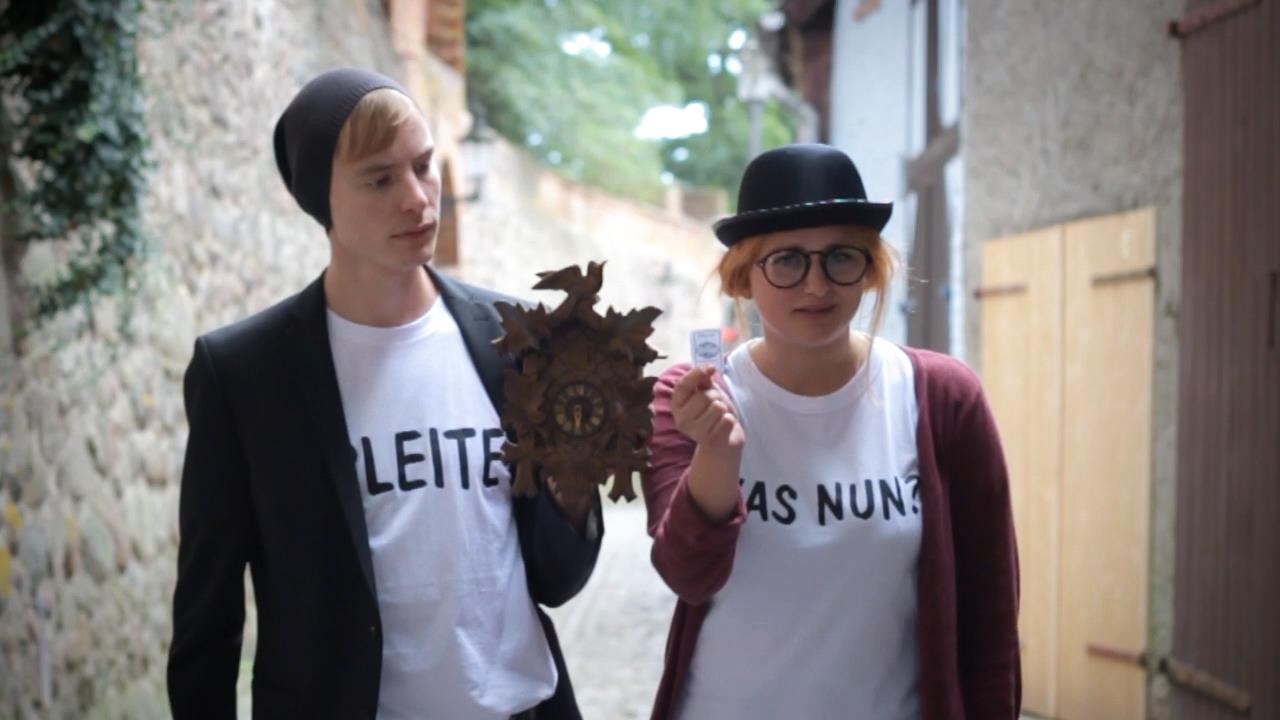 Zwei Personen tragen T-Shirts mit der Aufschrift "Pleite was nun". Sie stehen nebeneinander und lächeln in die Kamera.