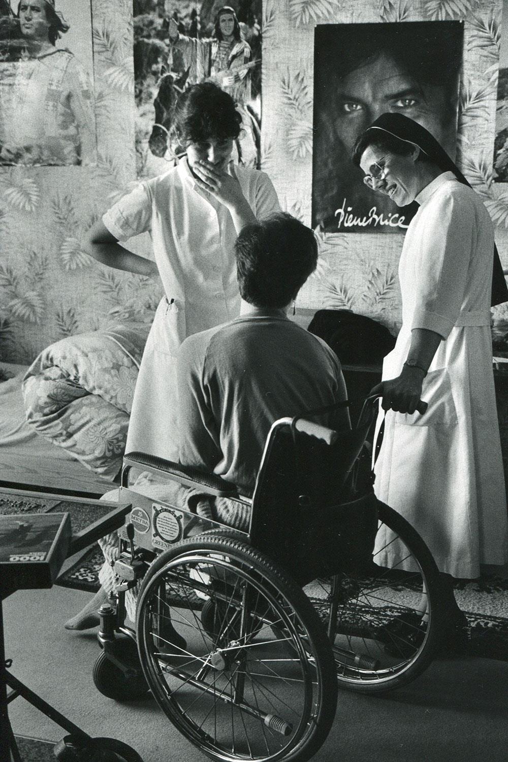 Junger Mensch sitzt in seinem Zimmer im Rollstuhl. Neben ihm stehen zwei Pflegerinnen. Eine davon trägt ein Ordensgewand.