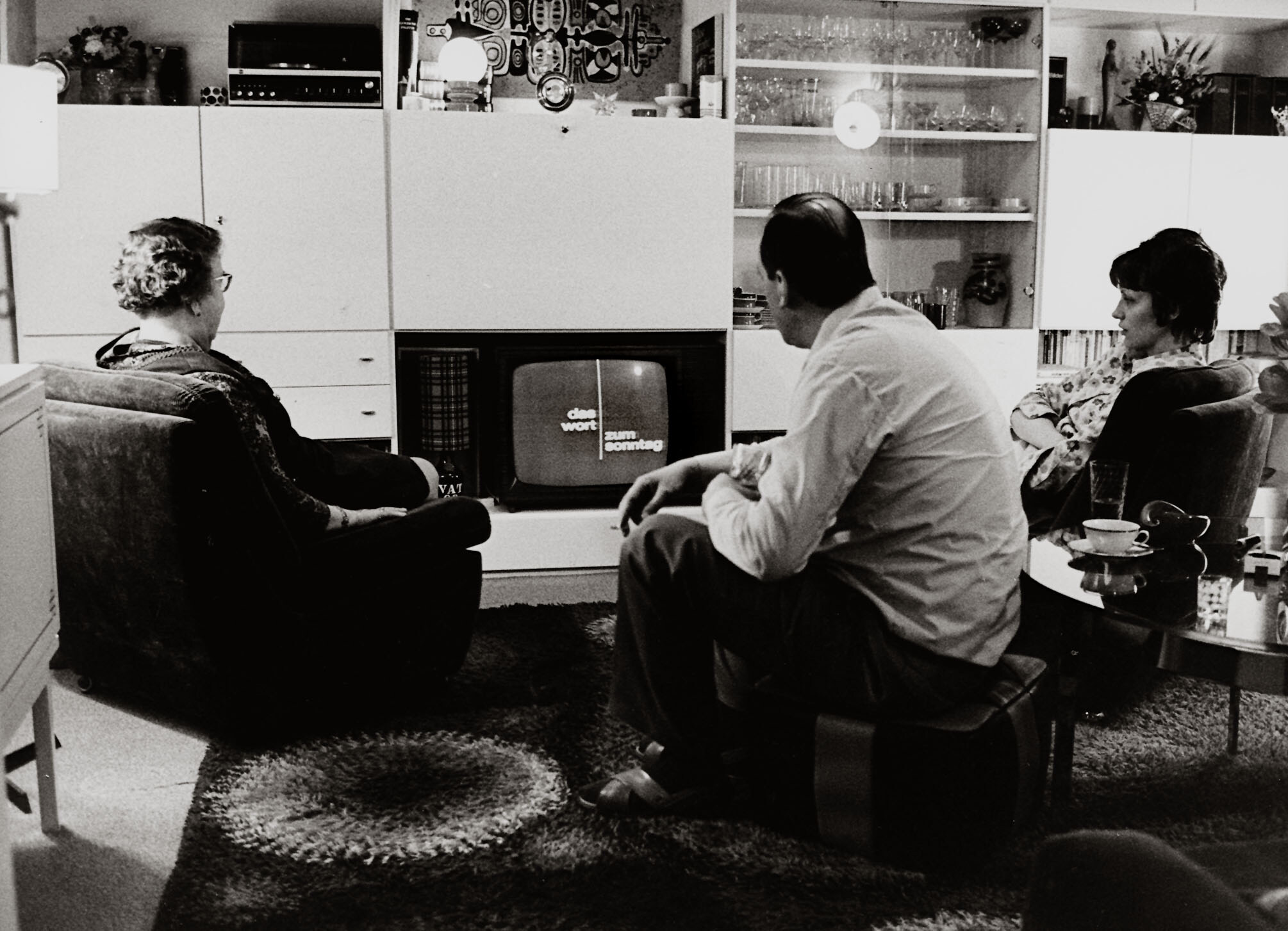 Drei Menschen sitzen mit Rücken zur Kamera in einem Raum und schauen Fernsehen.