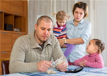 Ein Mann hat Euro-Scheine in der Hand und sitzt am Tisch. Eine Frau mit zwei Kindern auf dem Arm schaut ihn an.