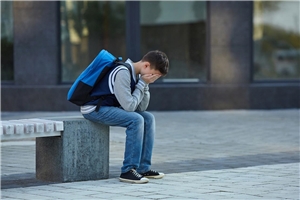 Ein Junge sitzt gebeugt auf einer Bank und hält seine Hände vor sein Gesicht.