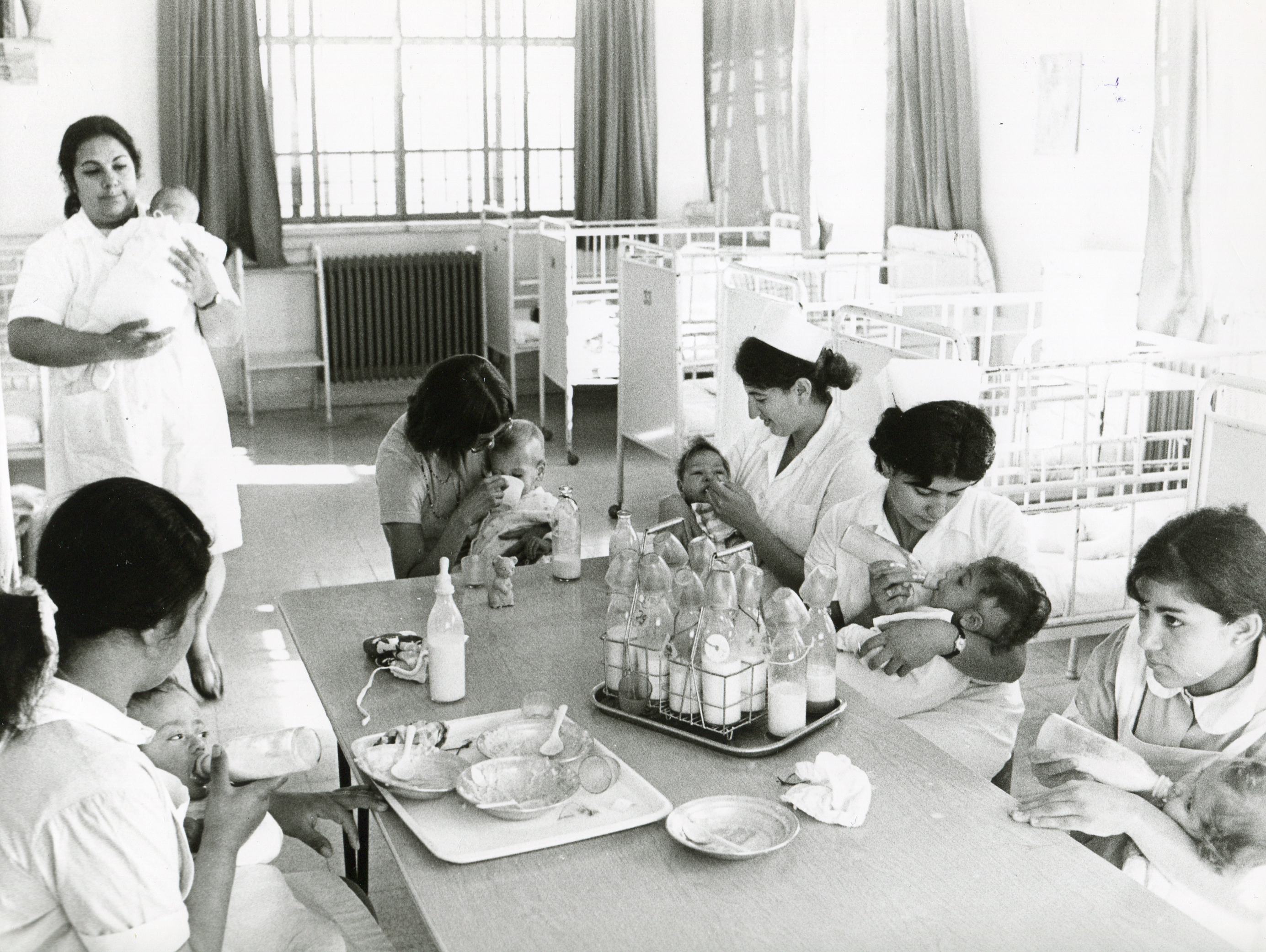 Fünf medizinische Fachkräfte im weiß bekleidet sitzen am Tisch mit Babies und geben essen,. Auf dem Tisch stehen zwei Tablets mit voller Flaschen. eine medizinische Fachkraft füttert hält ein Baby in der Hand und füttert.