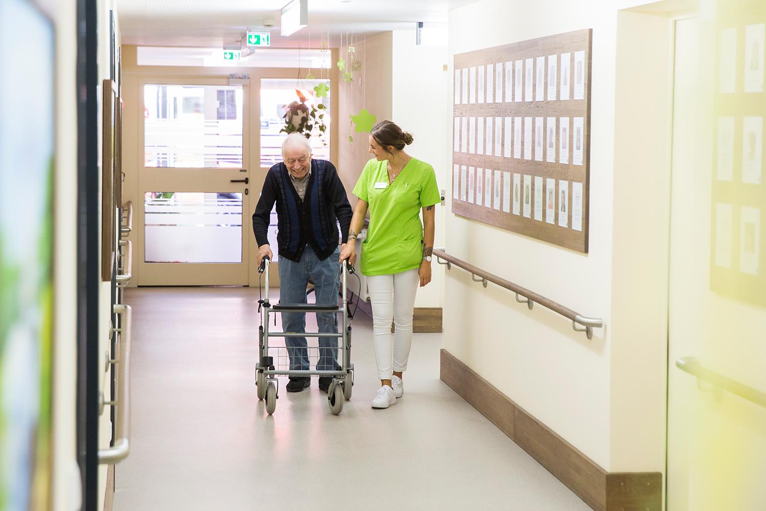 Eine Pflegerin läuft mit einem Mann mit Rollator durch den Gang eines Pflegeheims