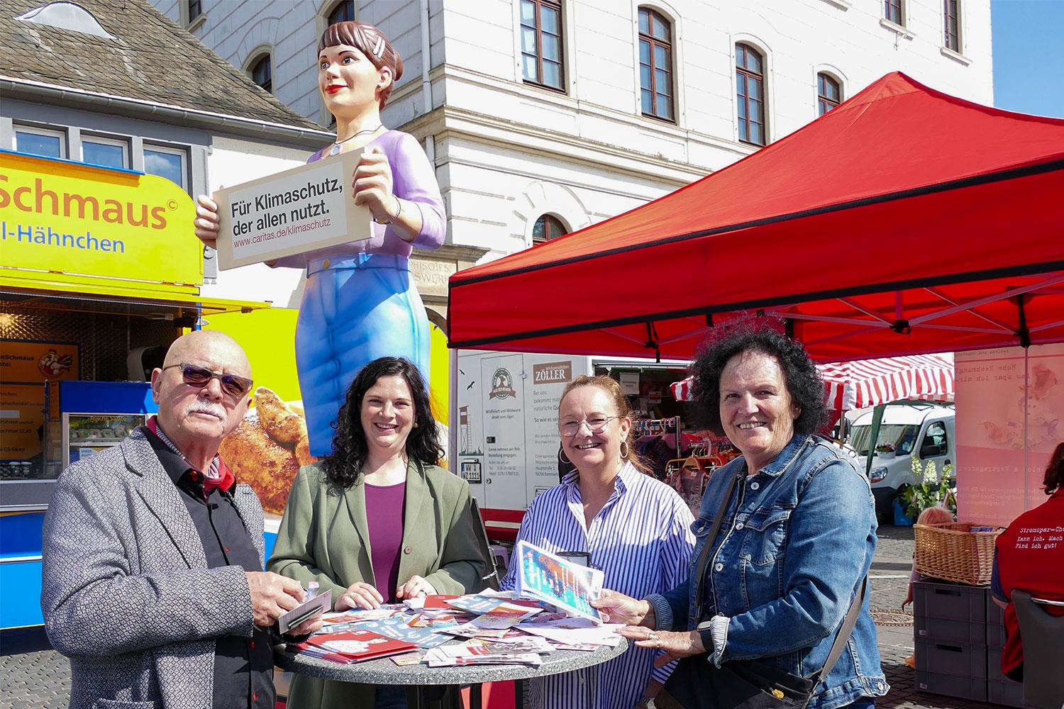 Die Jenny-Statue steht auf einem Wochenmarkt in Koblenz. Im Vordergrund unterhalten sich vier Personen.