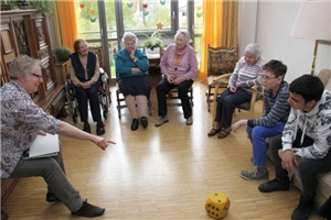 In einem offenen Stuhlkreis sitzen Senioren und Kinder. Rechts vorn liegt im Kreis ein gelber Ball aus Schaumstoff.