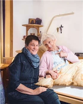 Eine Frau und eine bettlägige Seniorin halten lächelnd Händchen
