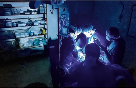Ein improvisierter OP-Raum mit einem Regal voller medizinischer Bedarfsartikel im Hintergrund - im Vordergrund ein 4-köpfiges OP-Team im Schein einer Handlampe.