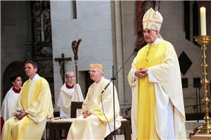 Auf dem Bild sitzen mehrere Geistliche, der Bischof steht am Mikrofon
