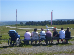 Die Gruppe sitzt auf einer Bank vor dem Flugplatz auf der Wasserkuppe. Alle schauen gespannt dem Flu