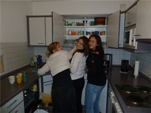 Drei junge Frauen betätigen sich in der Wohnküche. Sie ist an 3 Seiten mit Schränken und einer druch