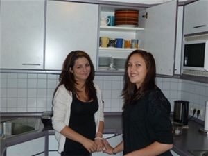 Zwei Frauen stehen in der Küche vor dem geöffneten Küchenschrank. Die beiden schauen uns lächelnd an